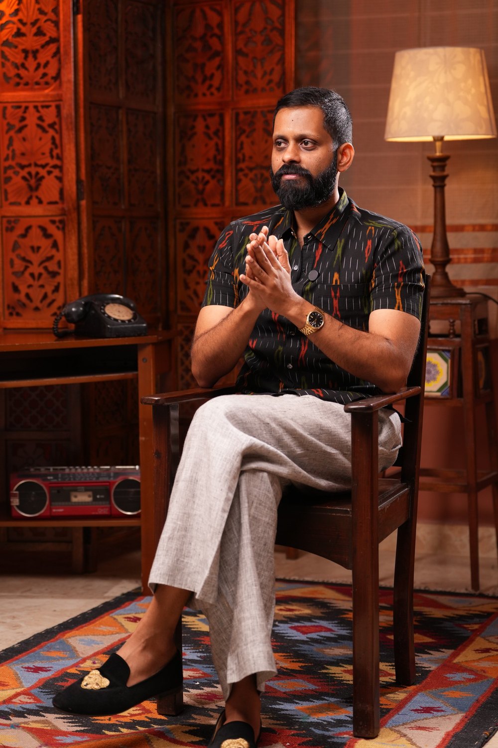 Shashank seated in a wooden chair, hands together, in a lamp-lit room.